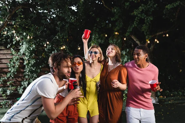 Homem cantando karaoke perto de amigos inter-raciais com copos de plástico ao ar livre — Fotografia de Stock