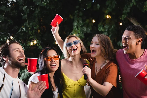 Mujeres multiétnicas con copas de plástico y micrófono cantando karaoke cerca de amigos - foto de stock