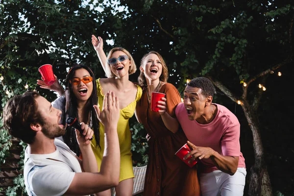 Man singing in microphone near positive multicultural friends with plastic cups outdoors in evening — Stock Photo