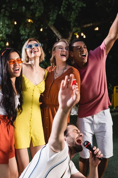 Межрасовые друзья с пластиковой чашкой поют рядом с мужчиной с микрофоном во время вечеринки на открытом воздухе — стоковое фото