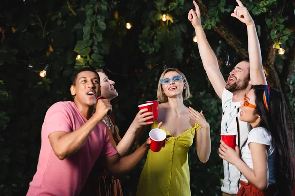 Africano americano homem cantando karaoke perto de amigos com copos de plástico ao ar livre — Fotografia de Stock