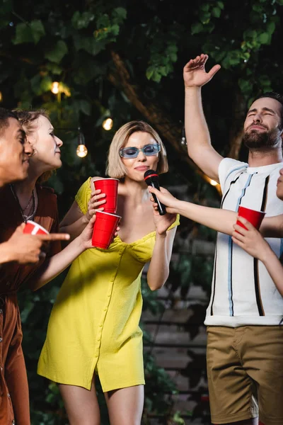 Young woman holding microphone near interracial friends with plastic cups outdoors — Stock Photo