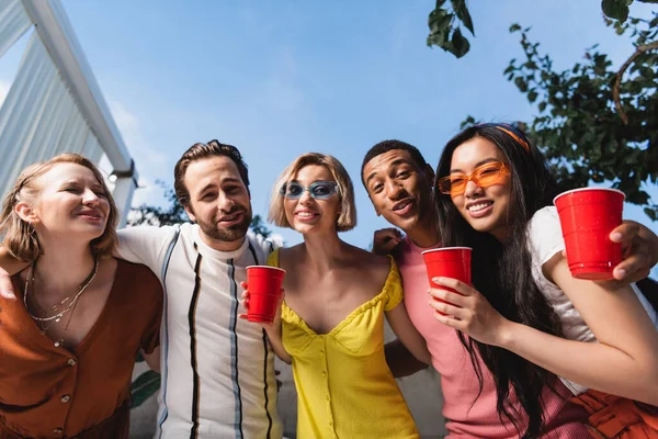 Vista ad angolo basso di amici multiculturali con drink guardando la fotocamera durante la festa — Foto stock