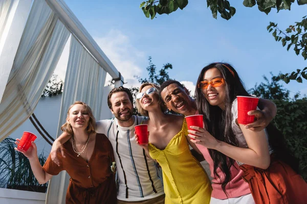 Feliz interracial de los jóvenes con vasos de plástico abrazando al aire libre — Stock Photo