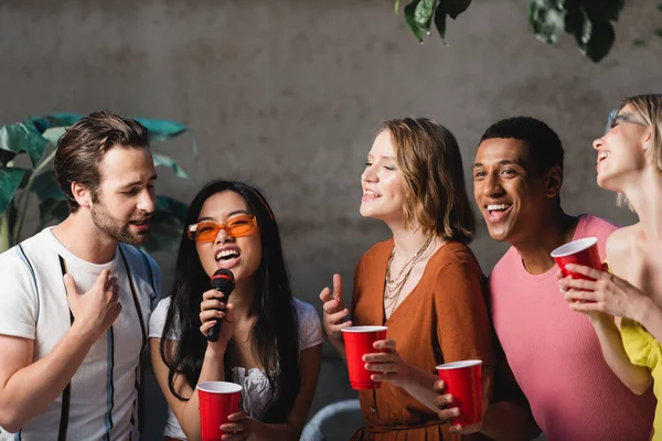 Asian woman with microphone singing near interracial friends holding plastic cups in evening — Stock Photo