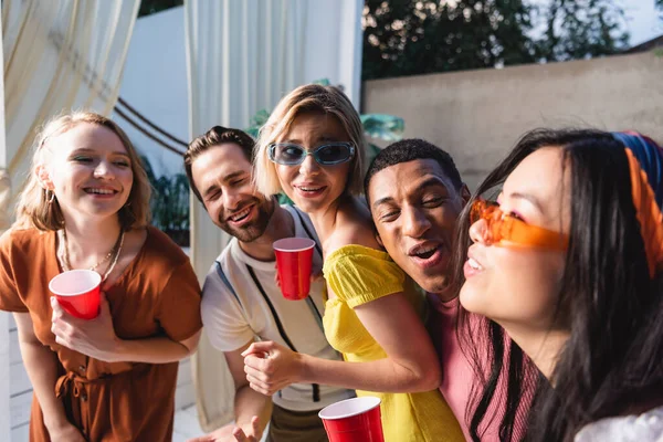 Amigos multiculturales con copas de plástico riendo y mirando a la mujer asiática al aire libre - foto de stock