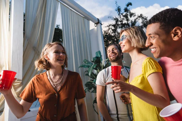 Donna sorridente con tazza di plastica che punta ad un amico vicino agli uomini interrazziali durante la festa — Foto stock