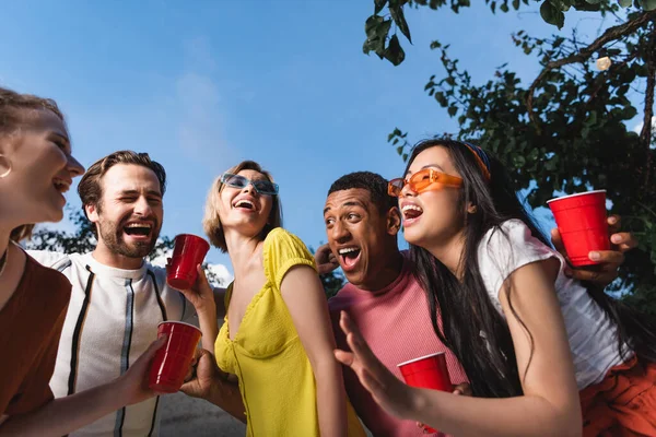 Vista de ángulo bajo de personas interracial excitadas con copas de plástico al aire libre - foto de stock