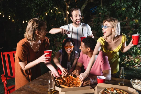 Jovens amigos multiétnicos tomando pizza perto de garrafas de cerveja ao ar livre — Fotografia de Stock