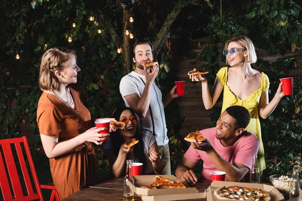 Felice multiculturale amici mangiare pizza vicino bicchieri di plastica e birra all'aperto — Foto stock