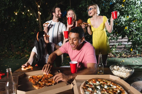 Africano americano tomando pizza perto de amigos sorridentes e cerveja ao ar livre — Fotografia de Stock