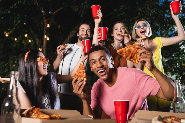 Homme afro-américain souriant tenant une pizza près d'amis flous avec des tasses en plastique à l'extérieur — Photo de stock