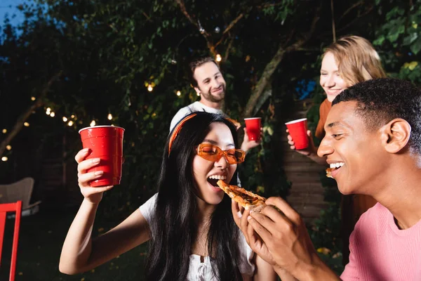 African american man feeding pizza to asian friend with plastic cup outdoors — Stock Photo