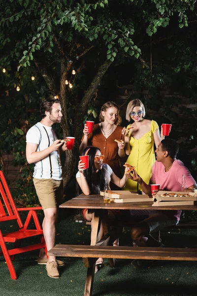 Amigos positivos con pizza mirando a un amigo afroamericano cerca de la cerveza bajo el árbol con guirnalda eléctrica - foto de stock