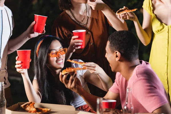 Felices amigos multiculturales sosteniendo vasos de plástico y trozos de pizza al aire libre - foto de stock