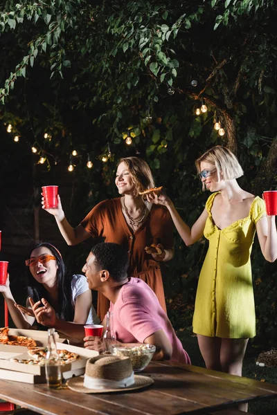 Межрасовые друзья с пиццей и пластиковыми стаканчиками проводят время на открытом воздухе во время вечеринки — стоковое фото