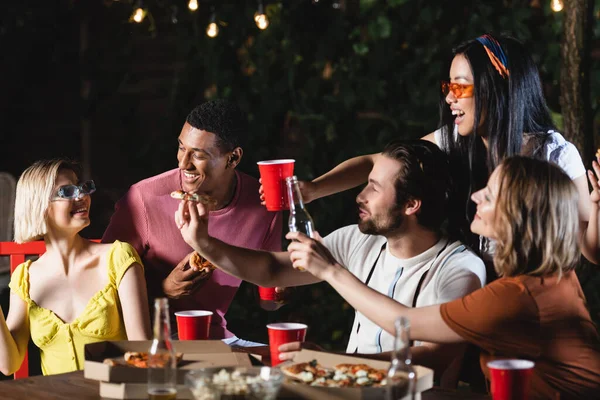 Sonrientes amigos interracial sosteniendo pizza cerca de la mujer y vasos de plástico en la mesa al aire libre - foto de stock