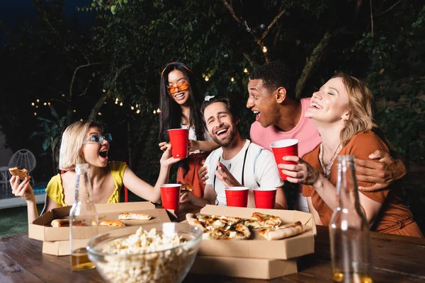 Emocionados amigos multiétnicos que pasan tiempo cerca de comida borrosa y botellas de cerveza al aire libre - foto de stock