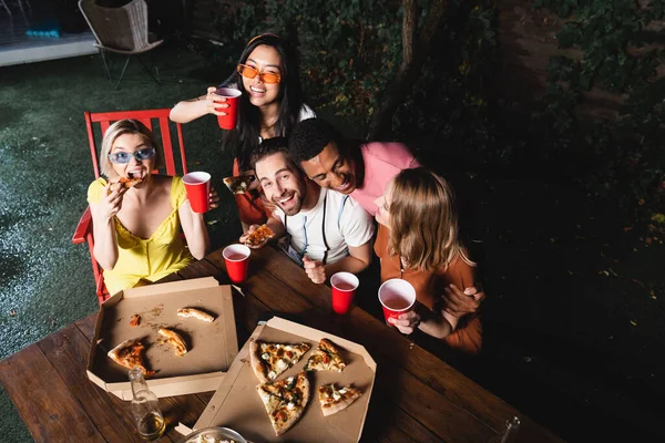 High angle view of positive interracial friends eating pizza and holding beer outdoors — Stock Photo