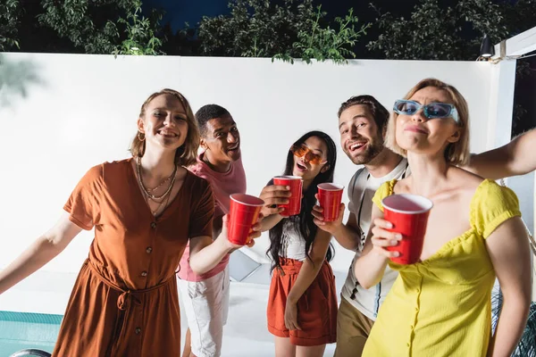 Interracial people with plastic cups looking at camera near swimming pool at night — Stock Photo