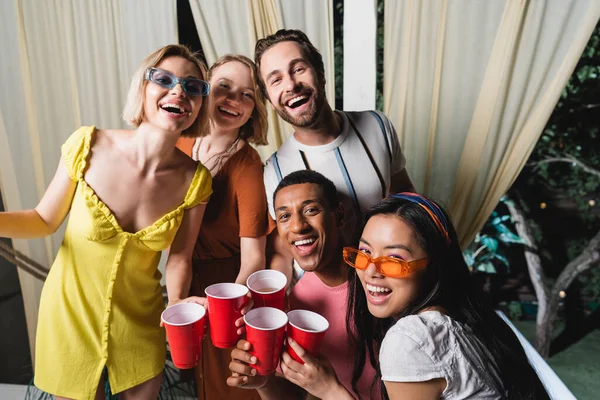 Молодые мультикультурные друзья с пластиковыми чашками, улыбающиеся в камеру во время летней вечеринки в патио — стоковое фото