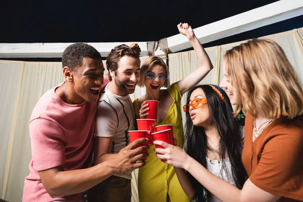 Excited multiethnic friends showing yes gesture and toasting with plastic cups in patio — Stock Photo