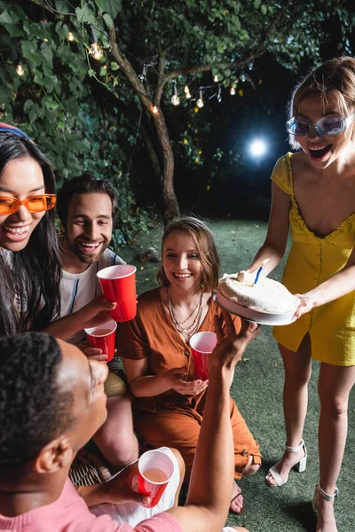 Woman holding birthday cake near interracial friends with plastic cups outdoors — Stock Photo