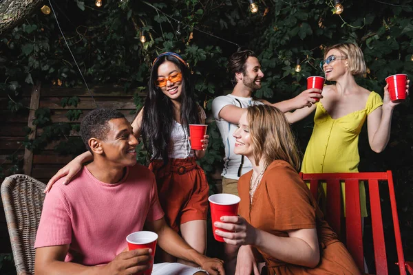 Young interracial friends with plastic cups talking during summer party in evening — Stock Photo