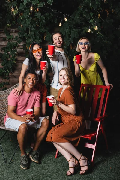 Sorrindo amigos multiétnicos com copos de plástico olhando para a câmera durante a festa — Fotografia de Stock