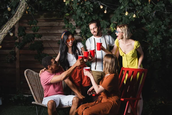 Fröhliche multikulturelle Freunde stoßen mit Plastikbechern unter Girlanden im Freien an — Stockfoto