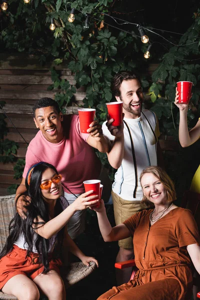 Улыбающаяся межрасовая молодежь с выпивкой в пластиковых чашках проводит время во время летней вечеринки — стоковое фото