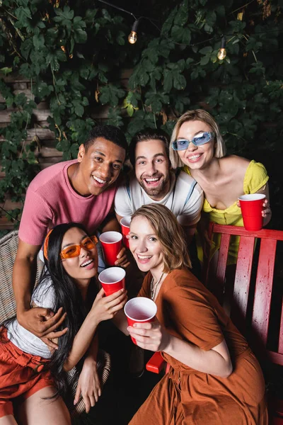 Smiling interracial friends with plastic cups hugging and looking at camera outdoors — Stock Photo