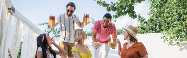 Smiling interracial men holding snacks and beer near friends in patio, banner — Stock Photo