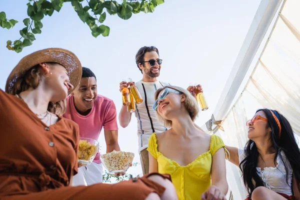 Visão de baixo ângulo de sorrir mulheres multiétnicas sentadas perto de amigos com cerveja e comida no pátio — Fotografia de Stock