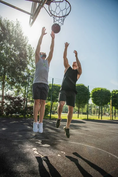 Young men jumping near ball and basketball hoop outdoors — Stock Photo