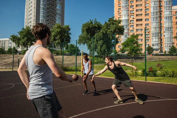 Interracial men playing basketball near blurred friend on playground — Stock Photo