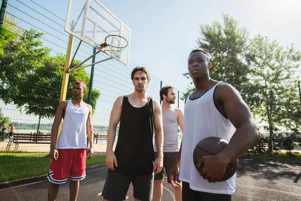 Jovens homens inter-raciais com bola de basquete olhando para a câmera no playground ao ar livre — Fotografia de Stock