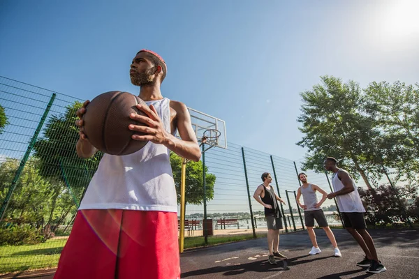 Vista basso angolo di uomo africano americano che tiene palla da basket vicino agli amici sul campo all'aperto — Foto stock