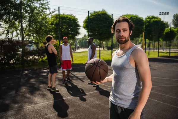 Young man holding basketball ball near blurred interracial friends on playground — Stock Photo