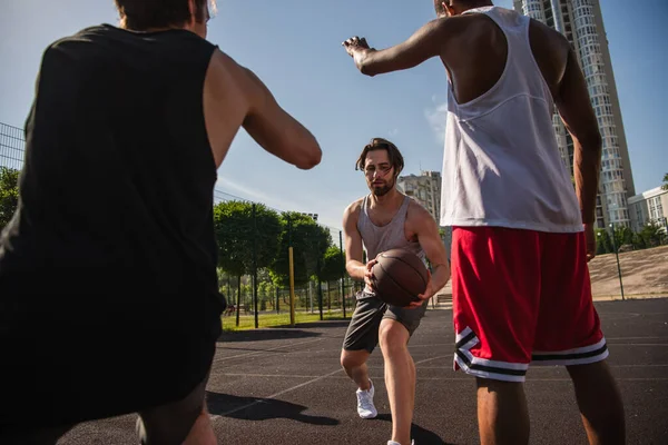 Joven sosteniendo pelota de baloncesto mientras juega con amigos multiétnicos al aire libre - foto de stock