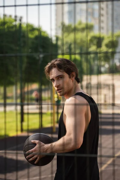 Hombre joven con pelota de baloncesto mirando la cámara cerca de la cerca al aire libre - foto de stock