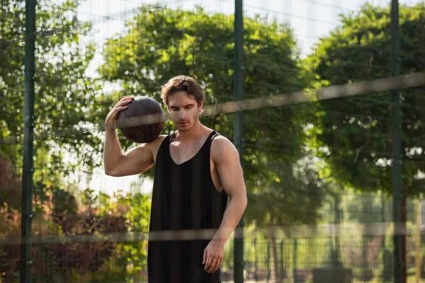 Esportista muscular segurando bola de basquete perto de cerca borrada ao ar livre — Fotografia de Stock