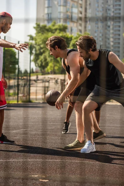 Vista lateral de hombres jóvenes jugando baloncesto cerca de amigo afroamericano al aire libre - foto de stock