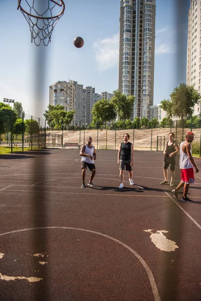 Interracial sportsmen looking at basketball ball near hoop on playground — Stock Photo