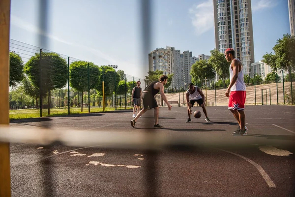 Hombres multiétnicos jugando baloncesto cerca de la cerca borrosa del patio de recreo - foto de stock