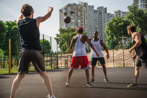 Interracial homens jogando basquete perto de amigo com as mãos levantadas em primeiro plano desfocado — Stock Photo