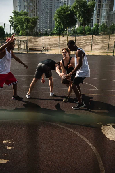 Возбужденный мужчина играет в баскетбол с многонациональными спортсменами на детской площадке — стоковое фото