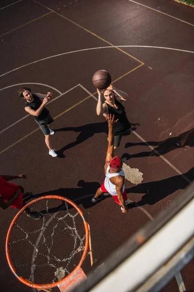 Vista aérea del hombre con pelota de baloncesto saltando cerca de amigos interracial y aro al aire libre - foto de stock