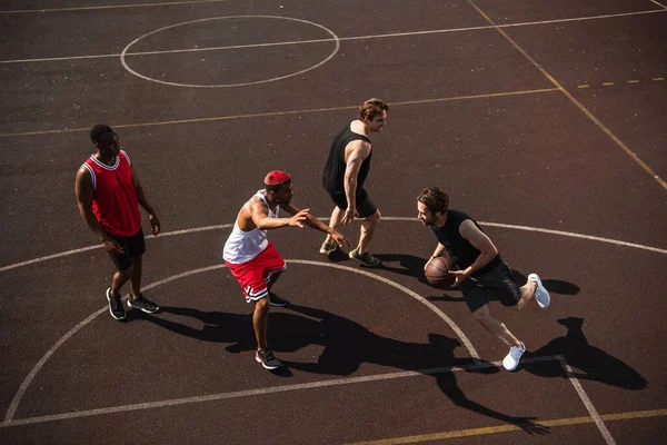 Vista ad alto angolo dell'uomo con pallone da basket in esecuzione sul parco giochi vicino agli amici multietnici — Foto stock