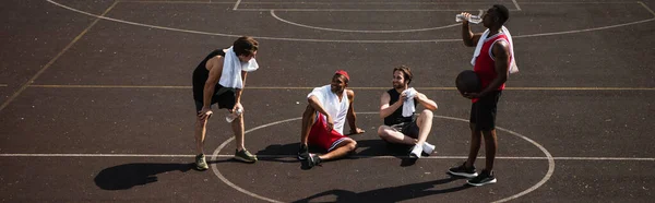 Hombres alegres interracial con pelota de baloncesto y toallas descansando en el patio de recreo, pancarta - foto de stock